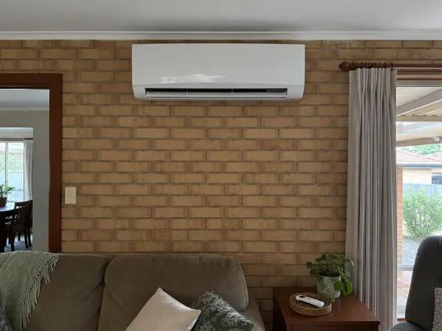 White split system mounted high on a brick wall in a living room with curtains and a sofa in Hillbank